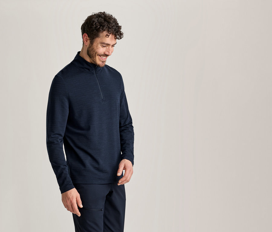 Un homme souriant aux cheveux bouclés porte un haut à manches longues bleu foncé avec un quart de zip et un pantalon bleu foncé, regardant vers le bas.