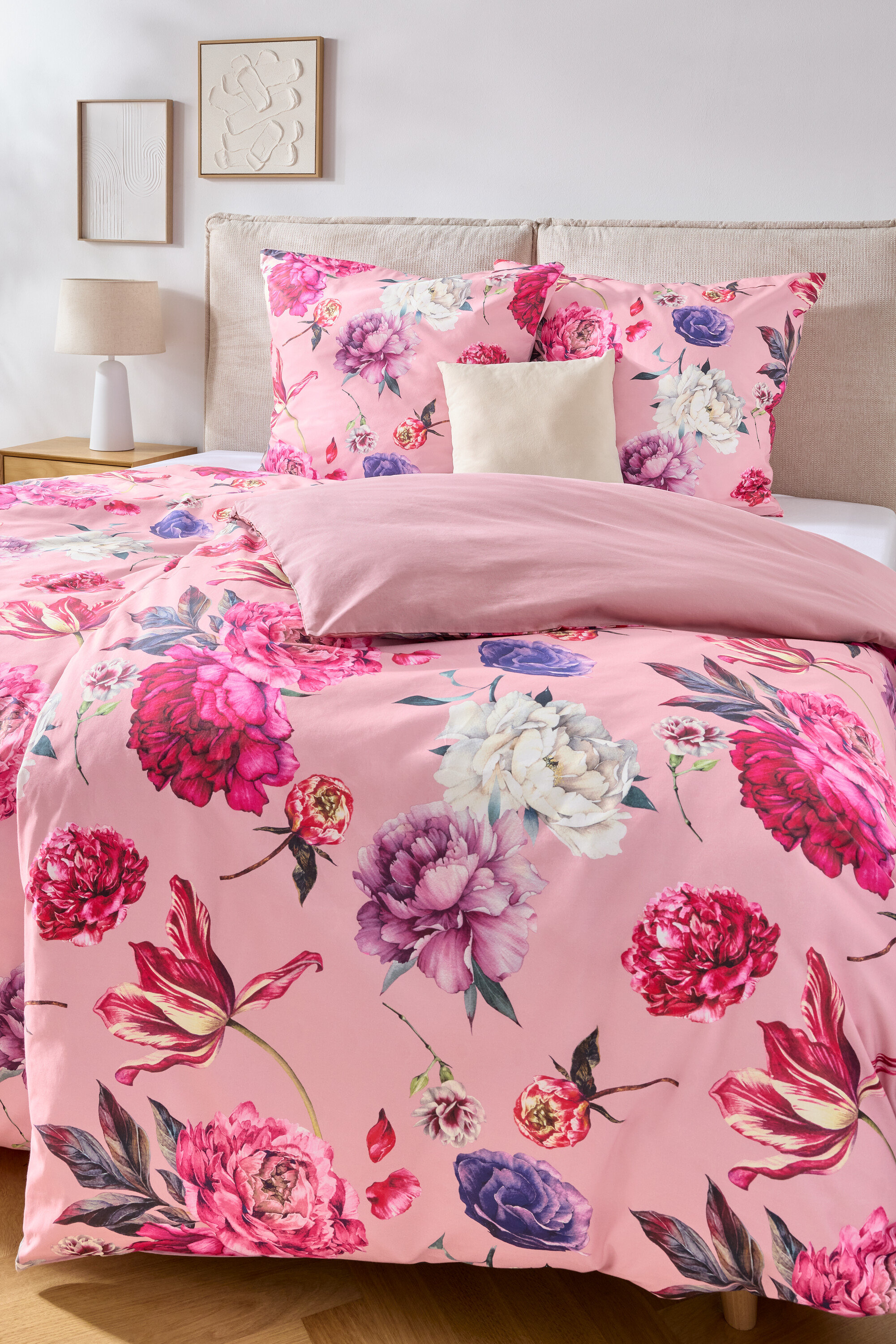 Gros plan d'un lit avec des draps roses à motif floral, une tête de lit beige et deux images encadrées au mur.