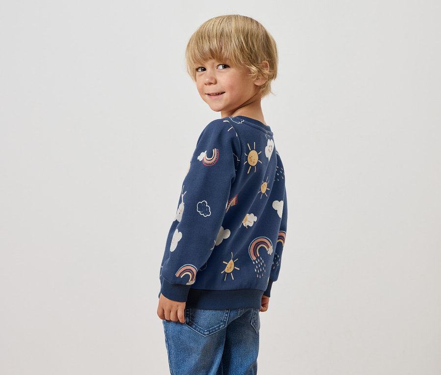 Un garçon aux cheveux blonds regarde par-dessus son épaule, portant un sweat-shirt bleu avec des motifs d'arc-en-ciel, de nuages et de soleil.