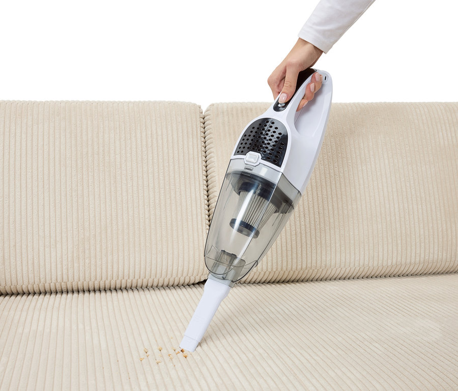 Une main aspire des miettes d'un canapé beige avec un aspirateur sans fil 2 en 1.