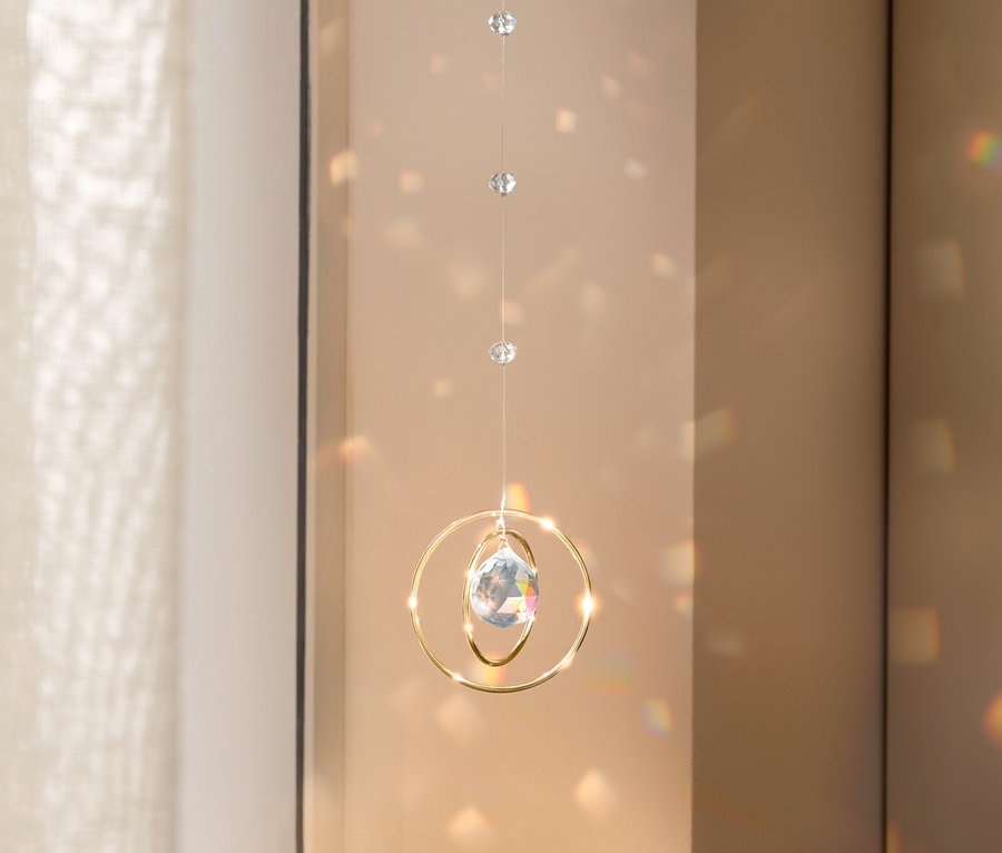 Un mobile en cristal avec des anneaux dorés est suspendu devant une fenêtre, projetant des taches de lumière sur le mur.