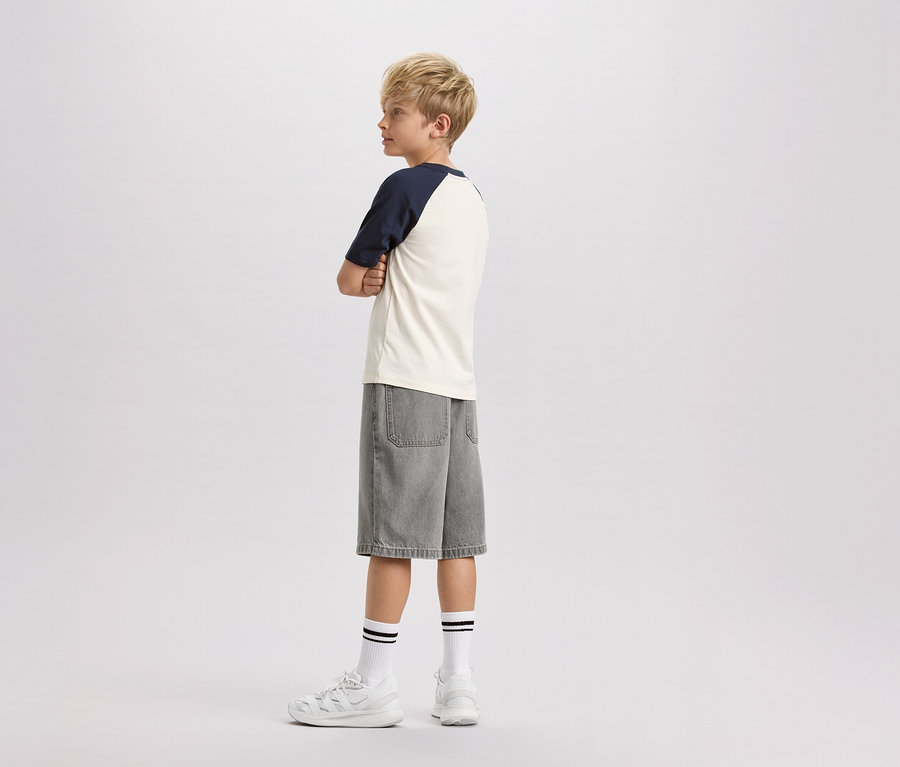 Un garçon blond se tient de profil, les bras croisés, portant un t-shirt crème à manches bleu marine, un short en jean gris, des chaussettes blanches à rayures noires et des baskets blanches.