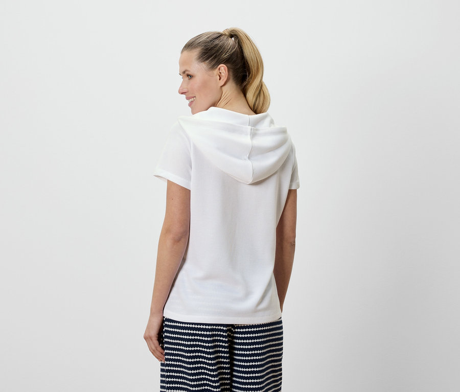 Une femme aux cheveux blonds porte un t-shirt à capuche blanc et un pantalon rayé bleu et blanc, vue de dos.
