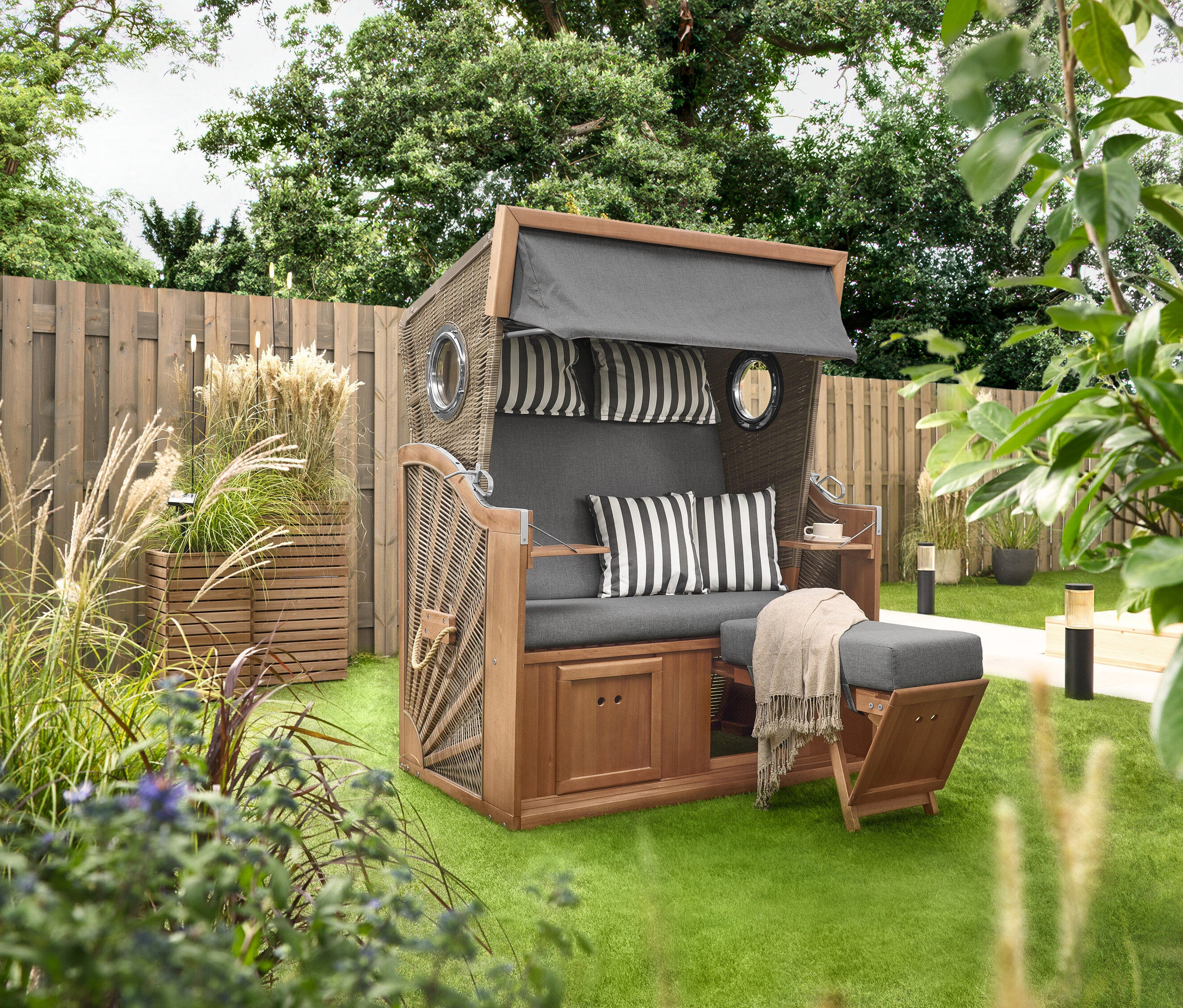 Un Premium-Strandkorb XL avec 2 tables pliantes est posé sur une pelouse, à côté de 2 colonnes à plantes « Tinus ». À l'arrière-plan, on voit 5 piquets à LED solaires.