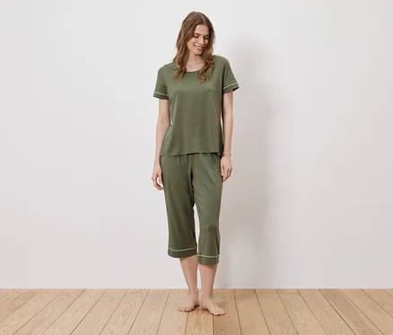 Une femme se tient pieds nus sur un plancher en bois et porte un pyjama vert olive avec des détails blancs.