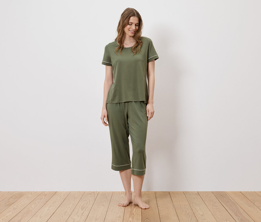 Une femme se tient pieds nus sur un plancher en bois et porte un pyjama vert olive avec des détails blancs.