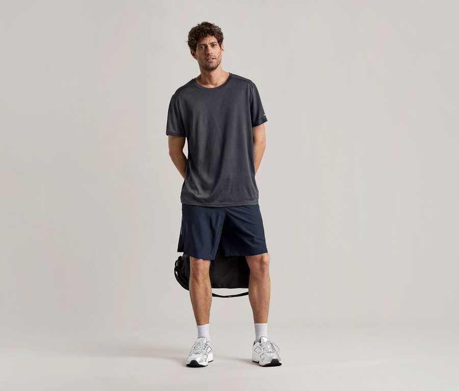 Un homme se tient debout sur un fond clair, portant un t-shirt de sport anthracite, un short et des baskets.