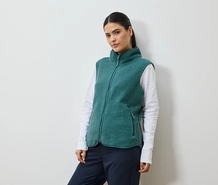 Femme avec queue de cheval portant un gilet en polaire vert sur un t-shirt blanc à manches longues et un pantalon bleu marine.