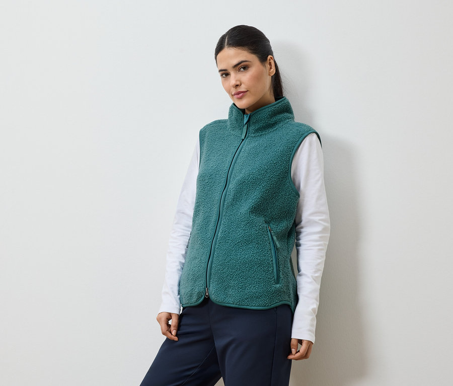 Femme avec queue de cheval portant un gilet en polaire vert sur un t-shirt blanc à manches longues et un pantalon bleu marine.
