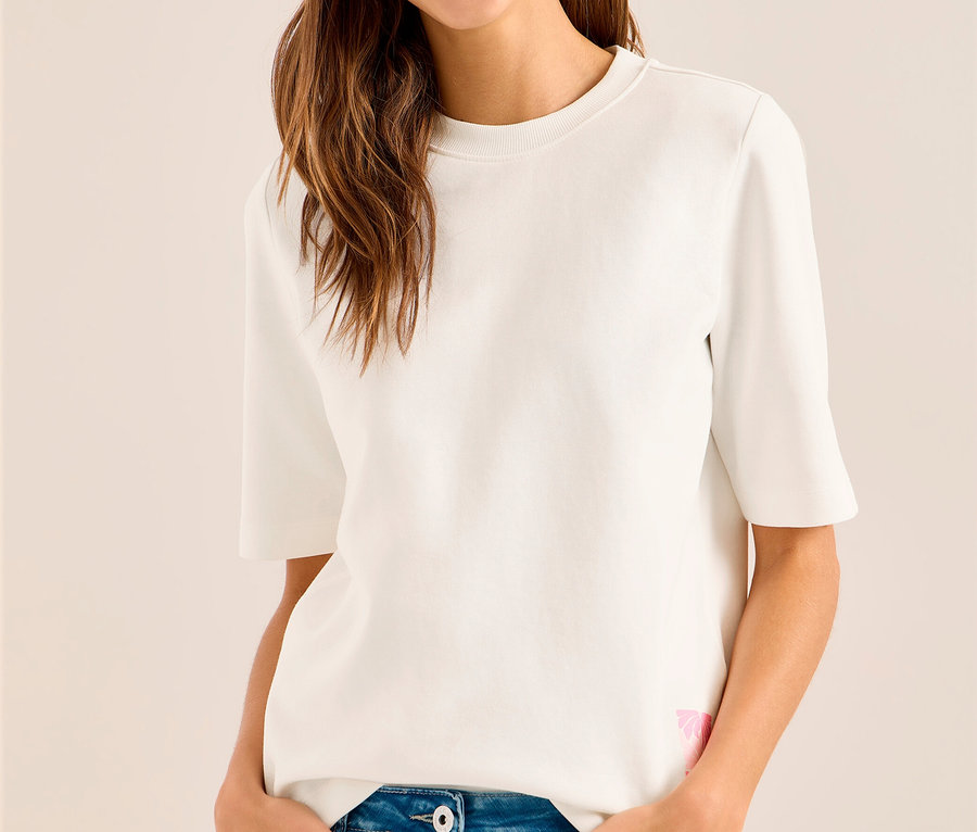 Femme souriante portant une casquette violette, un sweatshirt CECIL blanc et un jean bleu, les mains dans les poches.