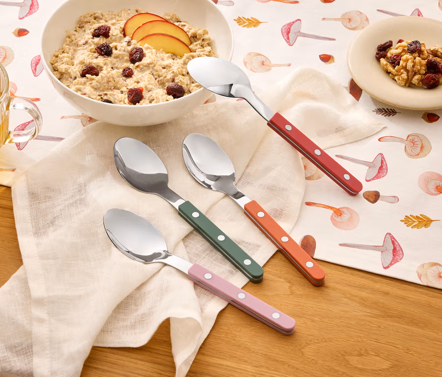 Sur une table à motifs de champignons se trouvent un bol de porridge, une tasse de thé, une assiette de noix et quatre cuillères à manches colorés.