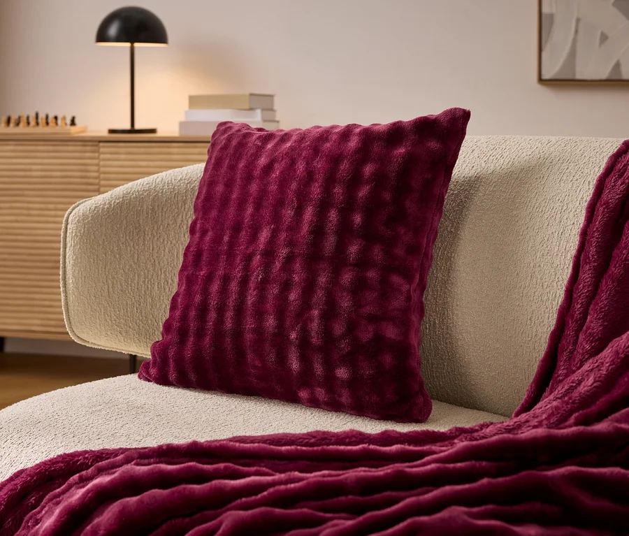 Coussin bordeaux sur un fauteuil crème, à côté d'une couverture bordeaux. En arrière-plan, une commode et une lampe.