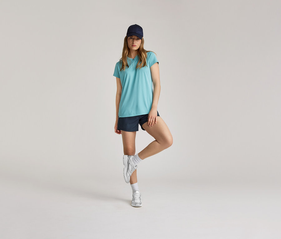 Une femme en t-shirt bleu, short et casquette se tient sur une jambe devant un fond clair.