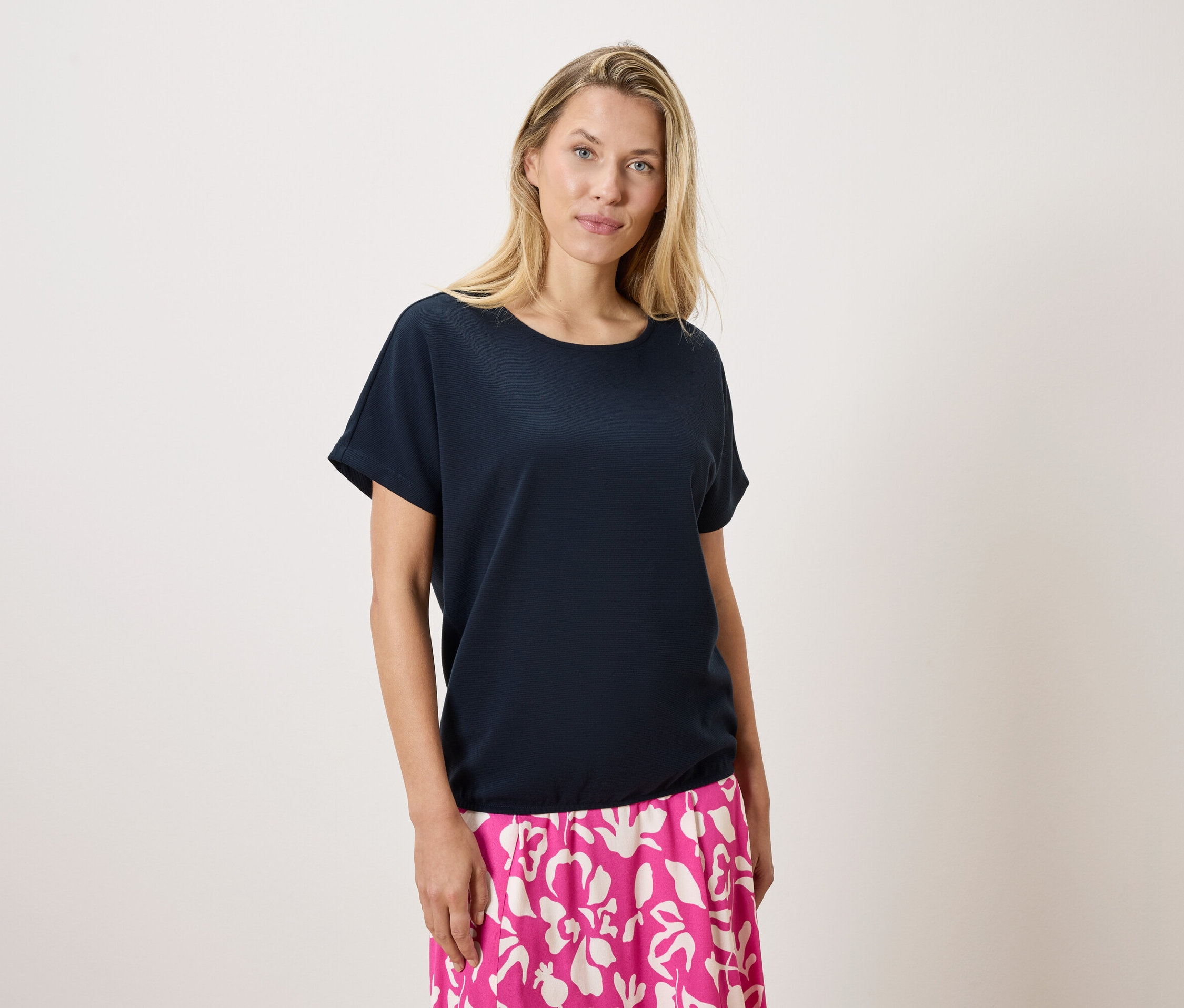 Une femme blonde porte un t-shirt bleu marine et une jupe rose et blanche à motifs sur un fond blanc.