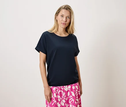 Une femme blonde porte un t-shirt bleu marine et une jupe rose et blanche à motifs sur un fond blanc.