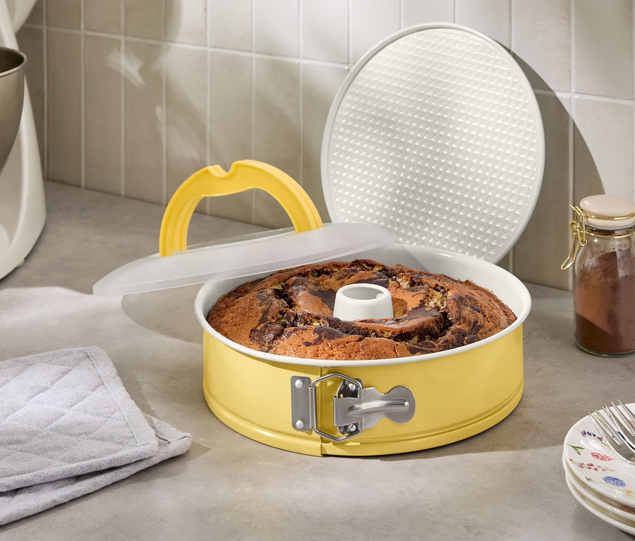 Gâteau marbré dans un moule à manqué jaune avec couvercle et poignée de levage à côté d'un robot culinaire et d'un pot à épices