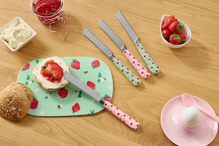 Sur une table en bois se trouvent un pain, une assiette à motifs de fraises, du fromage frais, de la confiture, des fraises et quatre couteaux à motifs de fraises.