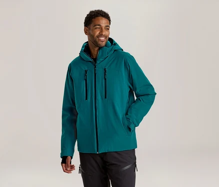 Un homme souriant aux cheveux bouclés foncés et barbu porte une veste à capuche sarcelle et un pantalon noir, regardant vers la droite avec une main dans sa poche.