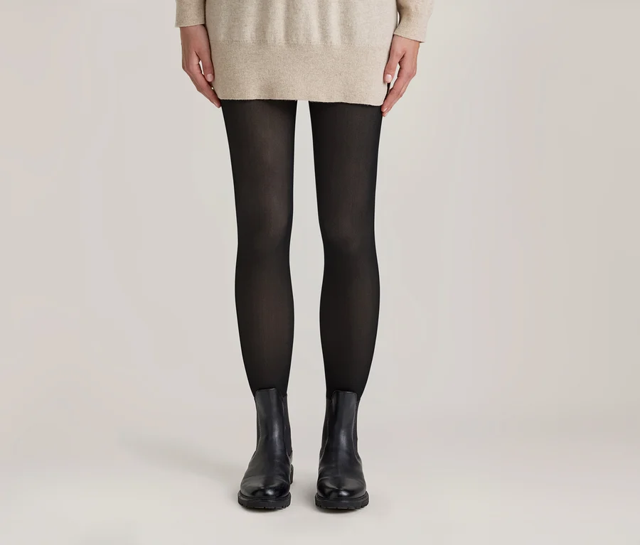 Personne debout portant un pull beige, des collants noirs et des bottines noires sur fond clair.