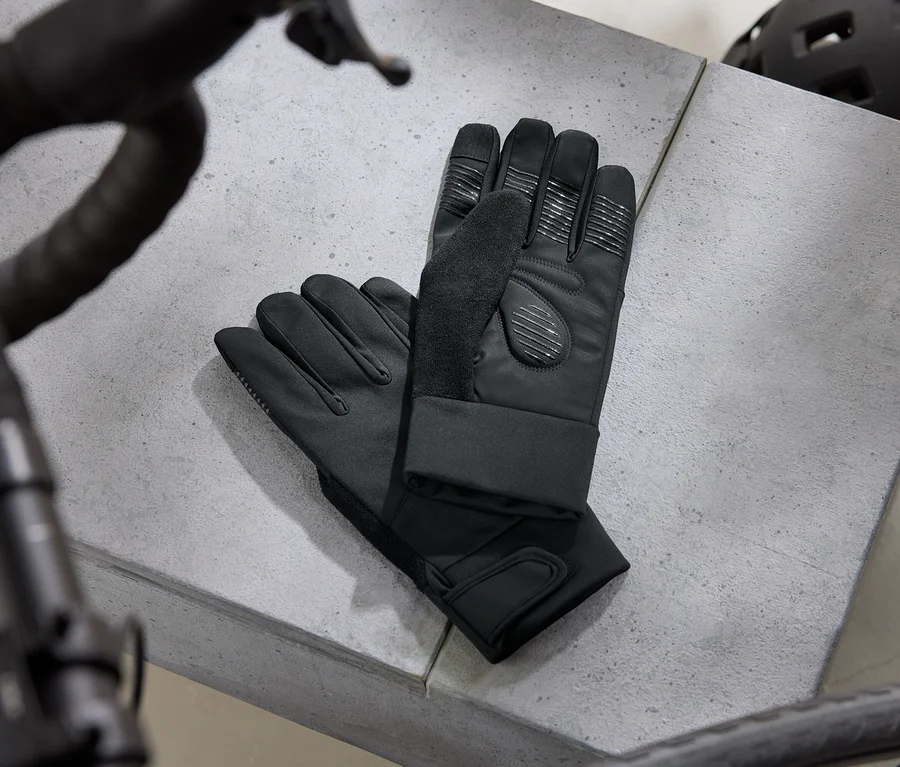 Une paire de gants de cyclisme noirs repose sur une surface en béton, avec un guidon de vélo à gauche et un casque à droite en arrière-plan.