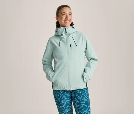 Femme souriante portant une veste à capuche vert menthe clair avec des fermetures éclair et des cordons noirs, et un pantalon bleu à motif géométrique blanc. Ses mains sont dans les poches de la veste.