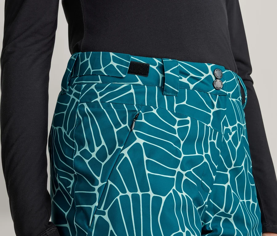 Gros plan d'une personne portant un haut noir à manches longues et un pantalon sarcelle avec un motif abstrait bleu clair. Le pantalon a une taille avec des boutons et une poche zippée.