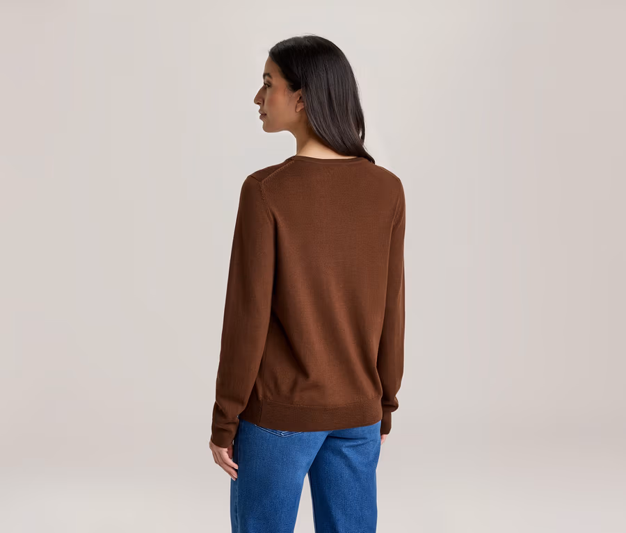 Une femme de dos, vêtue d'un pull marron à manches longues et d'un jean bleu. Sa tête est légèrement tournée vers la gauche.