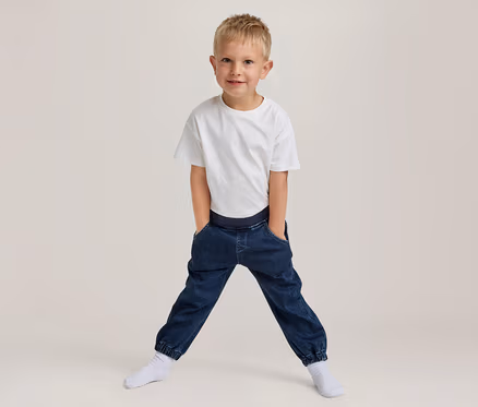 Jeune garçon blond souriant, portant un t-shirt blanc et un pantalon jogger bleu foncé avec les mains dans les poches, debout les jambes écartées en chaussettes blanches.