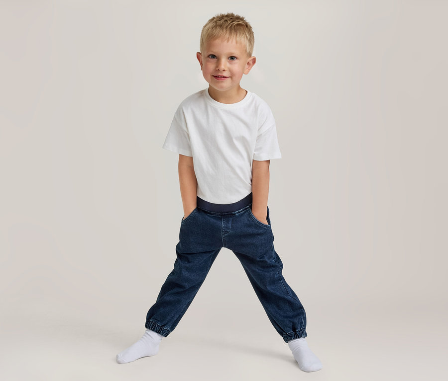 Jeune garçon blond souriant, portant un t-shirt blanc et un pantalon jogger bleu foncé avec les mains dans les poches, debout les jambes écartées en chaussettes blanches.