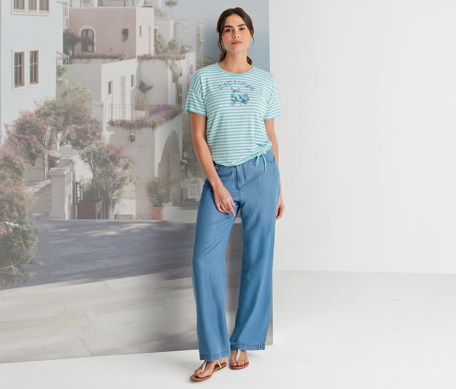 Une femme pose devant un paysage urbain méditerranéen, vêtue d'un T-shirt Olsen rayé avec un imprimé scooter, d'un pantalon large bleu clair et de sandales marron.