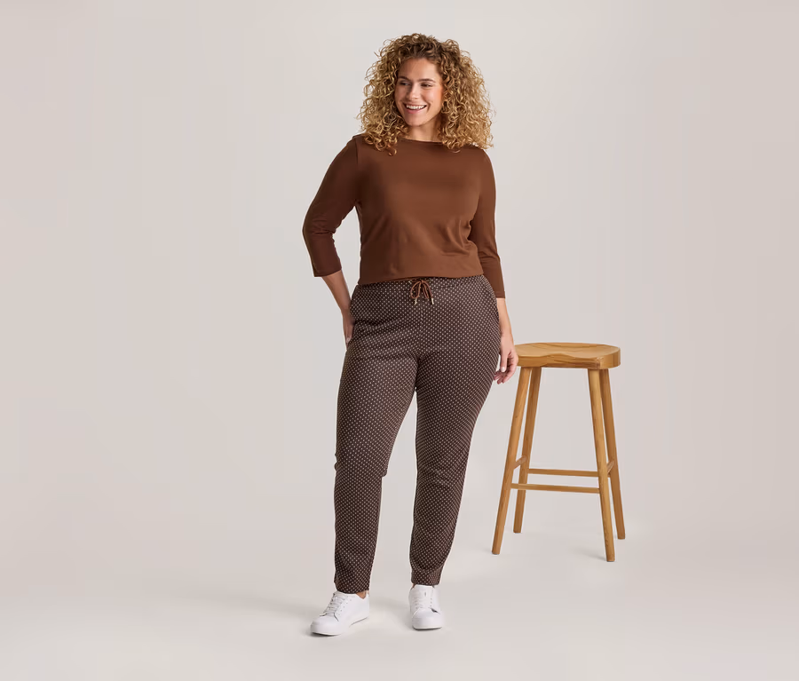 Femme souriante aux cheveux bouclés portant un haut marron à manches longues, un pantalon marron à motifs et des baskets blanches, debout à côté d'un tabouret en bois.