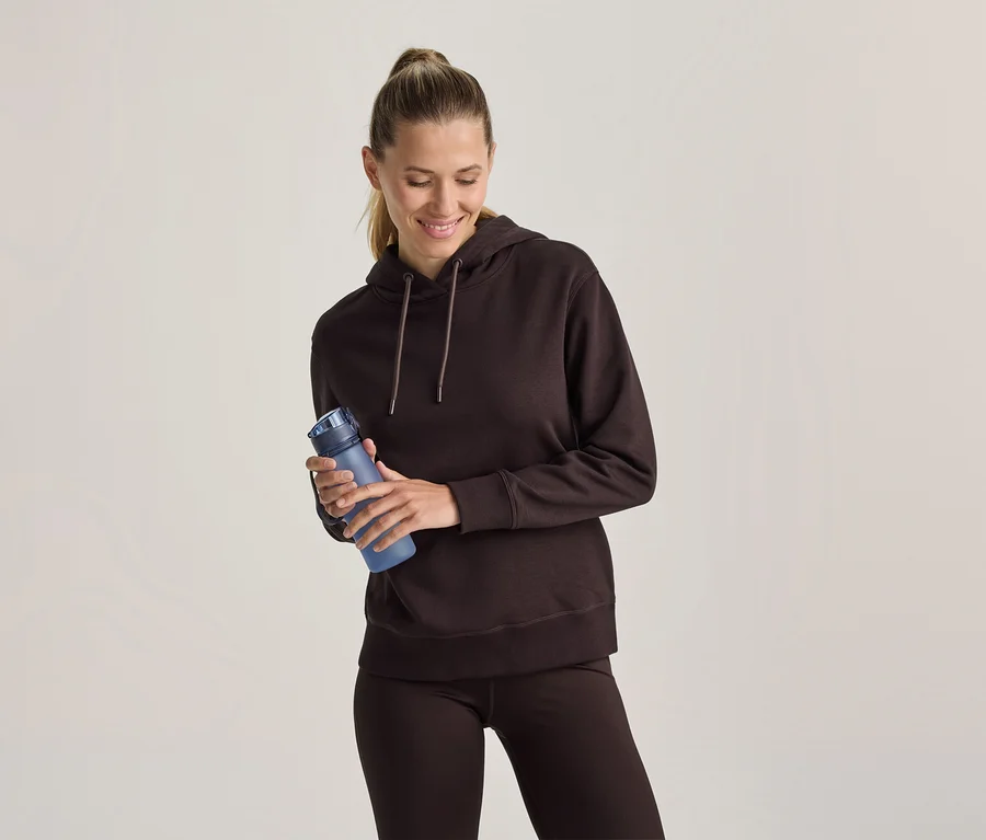 Femme en sweat à capuche marron et leggings marron tenant une bouteille d'eau bleue, souriant et regardant vers le bas.