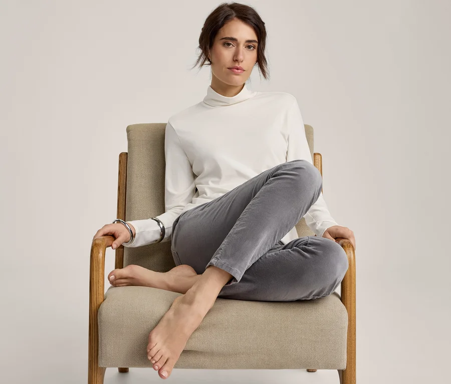 Femme assise pieds nus dans un fauteuil, vêtue d'un pull blanc à col roulé et d'un pantalon gris, les jambes croisées.