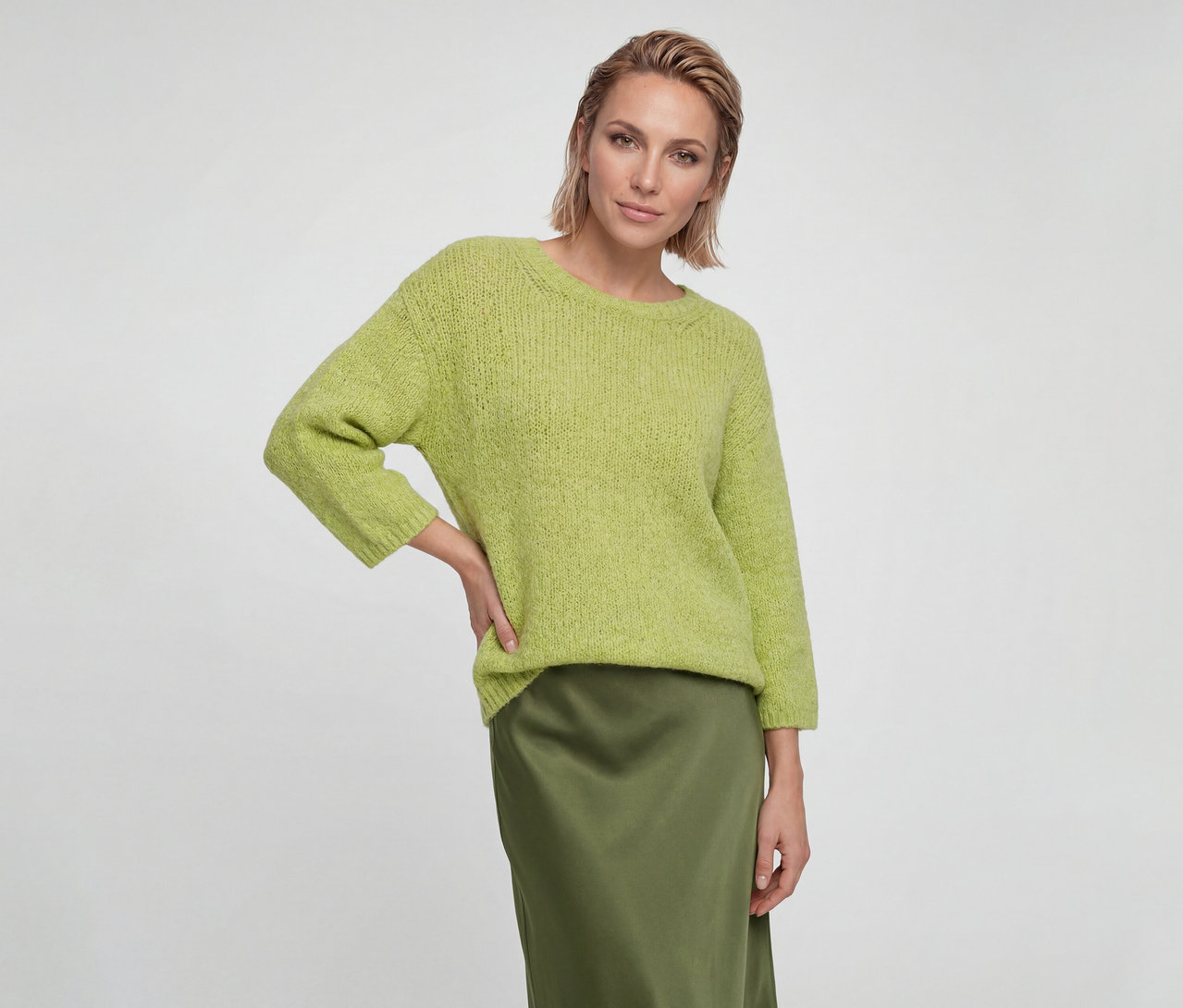 Femme aux cheveux blonds courts portant un pull en maille vert clair et une jupe en satin vert foncé, la main sur la hanche.