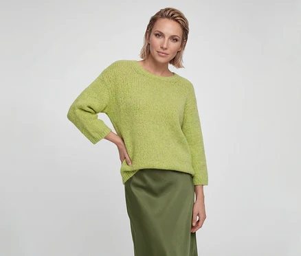 Femme aux cheveux blonds courts portant un pull en maille vert clair et une jupe en satin vert foncé, la main sur la hanche.