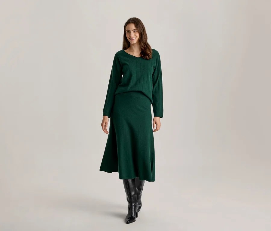 Femme souriante debout, portant un pull en maille vert foncé à col en V, une jupe midi assortie et des bottes noires montantes.