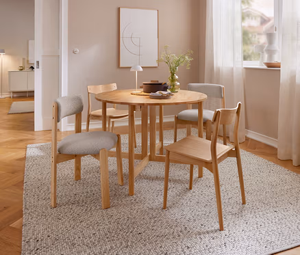 Une salle à manger avec une table ronde en bois et quatre chaises en bois sur un tapis clair. Sur la table se trouvent une lampe de table à LED sans fil beige, un vase de plantes et une casserole. À l'arrière-plan, une fenêtre avec des rideaux et un tableau sont visibles.