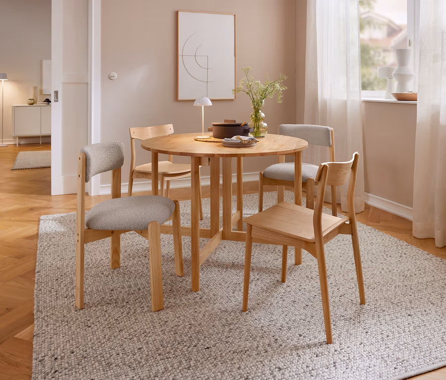 Une salle à manger avec une table ronde en bois et quatre chaises en bois sur un tapis clair. Sur la table se trouvent une lampe de table à LED sans fil beige, un vase de plantes et une casserole. À l'arrière-plan, une fenêtre avec des rideaux et un tableau sont visibles.