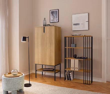 Un salon avec un parquet en bois clair et un tapis beige, présentant une armoire brun clair, une étagère avec des barres métalliques noires et un tabouret bleu avec un plateau en bois. Une lampe sur pied noire se tient à côté, et une fenêtre avec des rideaux est visible à gauche.
