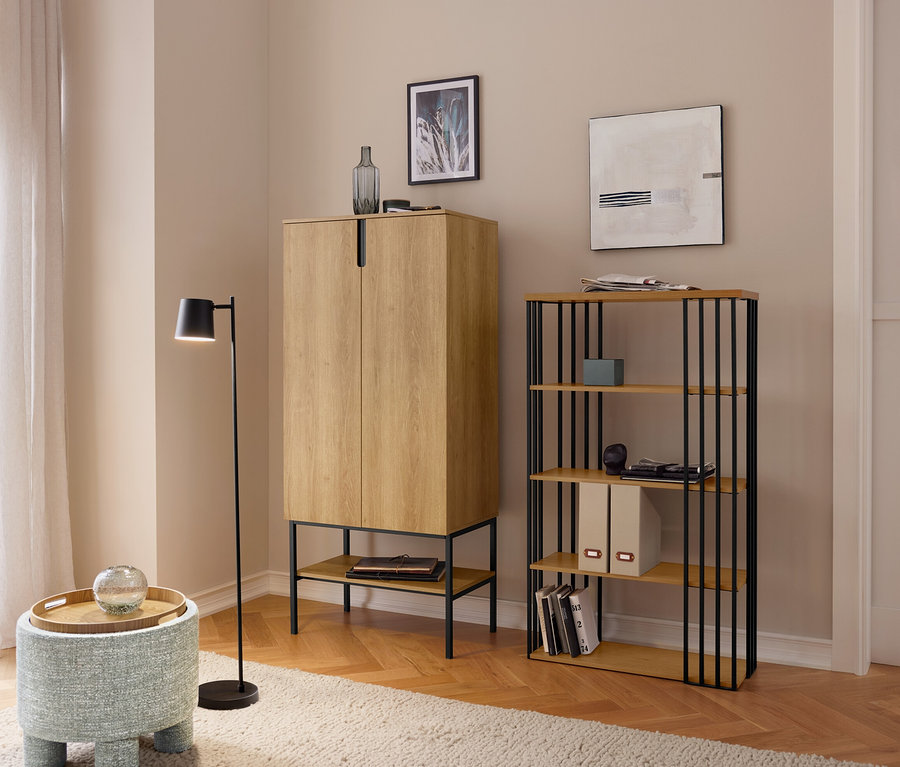 Un salon avec un parquet en bois clair et un tapis beige, présentant une armoire brun clair, une étagère avec des barres métalliques noires et un tabouret bleu avec un plateau en bois. Une lampe sur pied noire se tient à côté, et une fenêtre avec des rideaux est visible à gauche.