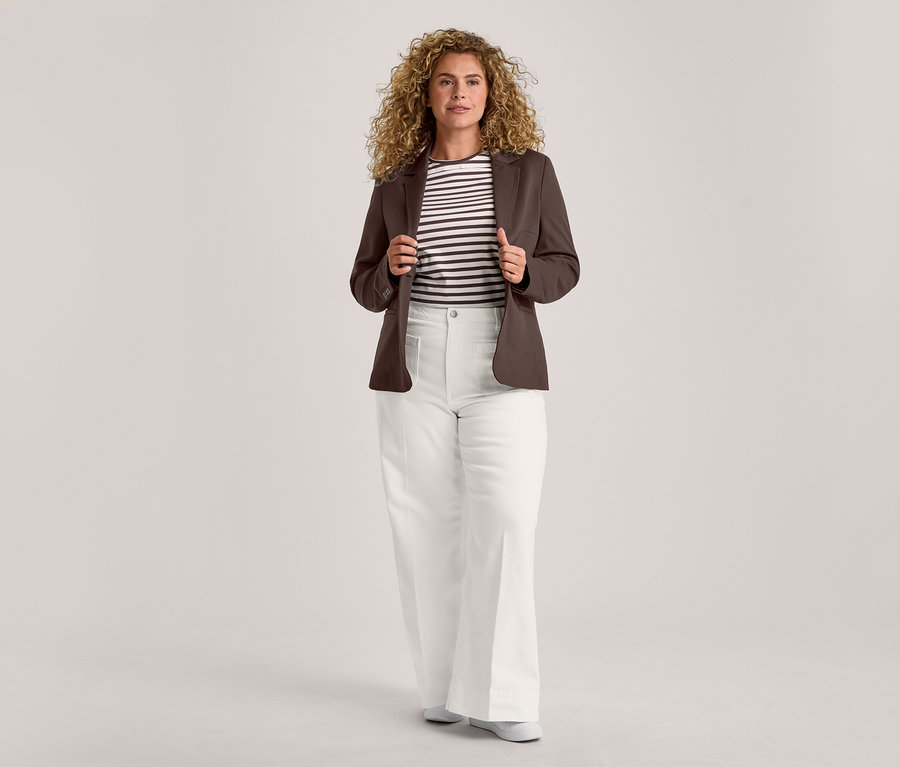 Une femme aux cheveux bouclés se tient de face, tenant les revers de son blazer marron ouvert. Elle porte un t-shirt rayé noir et blanc, un pantalon blanc à jambes larges et des chaussures plates de couleur claire.