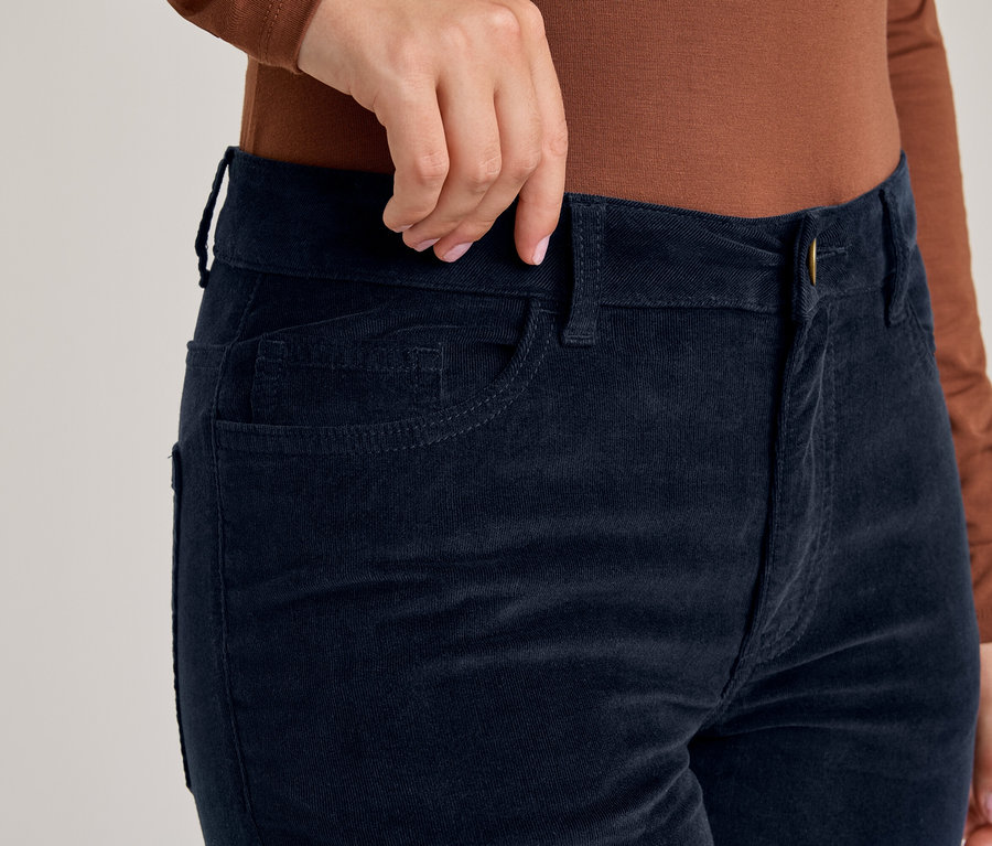 Main ajustant la taille d'un pantalon en velours côtelé bleu foncé, porté avec un t-shirt marron à manches longues.
