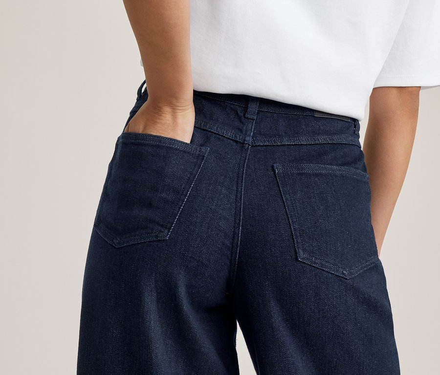 Vue de dos d'une personne portant un jean bleu foncé et un t-shirt blanc, une main dans la poche arrière.