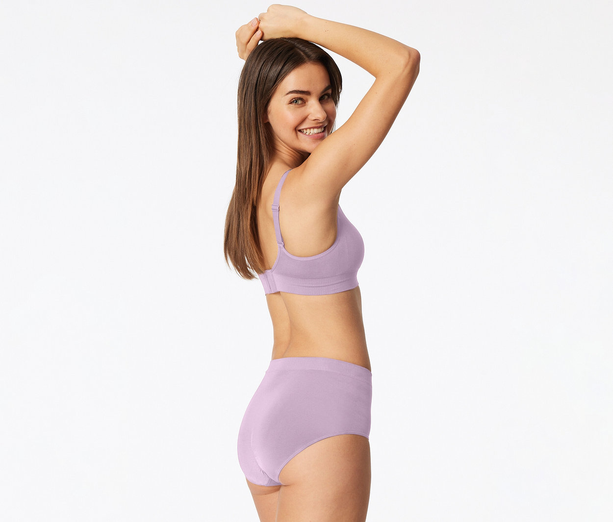 Une femme souriante portant un soutien-gorge et une culotte assortis de couleur lilas clair, regardant par-dessus son épaule, les bras levés.
