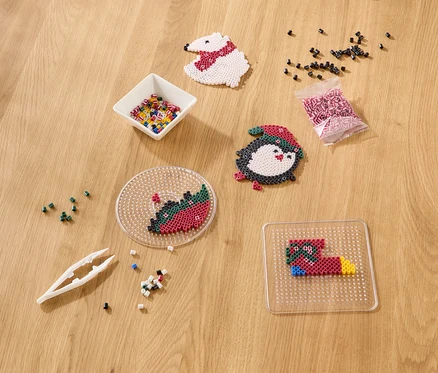 Matériel de bricolage sur une table en bois, comprenant des perles à repasser dans un bol et un sachet, une pince à épiler, des créations finies (ours polaire, pingouin) et inachevées sur des plaques.