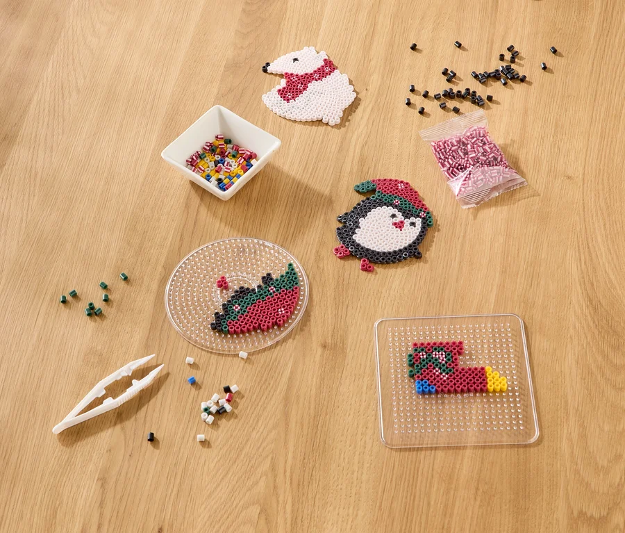 Matériel de bricolage sur une table en bois, comprenant des perles à repasser dans un bol et un sachet, une pince à épiler, des créations finies (ours polaire, pingouin) et inachevées sur des plaques.