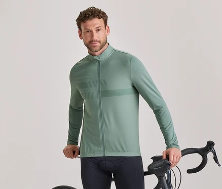 Un homme aux cheveux bruns courts et bouclés et à la barbe légère, portant une veste de cyclisme à manches longues vert sauge avec un motif subtil et un cuissard noir, se tient debout, tenant le guidon d'un vélo de route noir et regardant l'appareil photo.