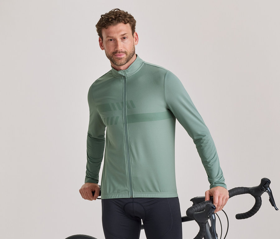 Un homme aux cheveux bruns courts et bouclés et à la barbe légère, portant une veste de cyclisme à manches longues vert sauge avec un motif subtil et un cuissard noir, se tient debout, tenant le guidon d'un vélo de route noir et regardant l'appareil photo.