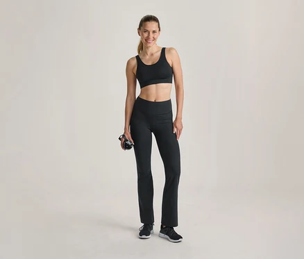 Femme souriante en soutien-gorge de sport noir et legging évasé noir, tenant une bouteille d'eau noire, debout sur un fond clair.