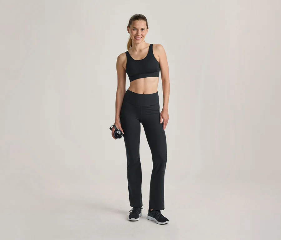 Femme souriante en soutien-gorge de sport noir et legging évasé noir, tenant une bouteille d'eau noire, debout sur un fond clair.